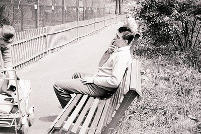 A young man in a sweater and slacks sits pensively on a wooden bench. A partial view of a woman and a vintage scooter are vis...