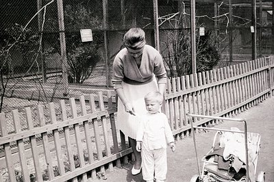 A mother and young boy stand near a picket fence; she appears to be adjusting his clothing. The scene, likely a zoo or park, ...