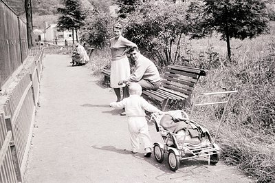 A young boy pulls a vintage baby stroller along a paved path. A couple stands nearby, observing him. A seated person is visib...