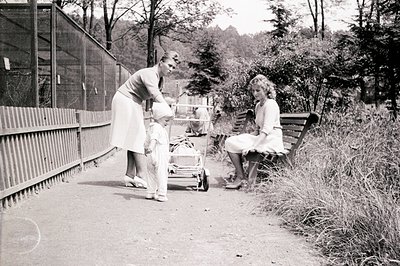 A young child, likely a toddler, stands near a vintage pram on a paved path. Two women, possibly a mother and nanny, are pres...
