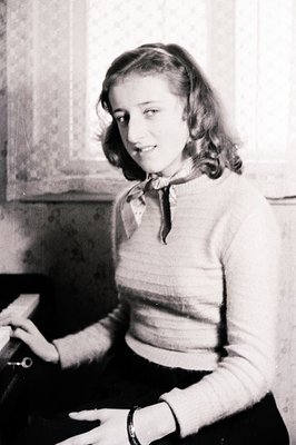 Young woman in a ribbed sweater and dark skirt, seated with a slight smile. A patterned scarf adorns her neck. The background...