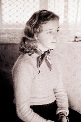 A young woman with styled hair and a dark scarf sits indoors, captured in a medium shot. She's wearing a textured sweater and...