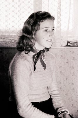 Young woman in a textured sweater and dark skirt, a silk scarf knotted at her neck. Seated indoors, looking over her shoulder...