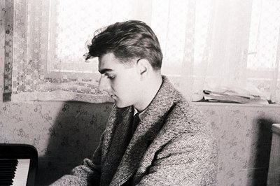 A young man in a textured overcoat sits near a window, appearing contemplative. The simple interior shows a patterned wall an...
