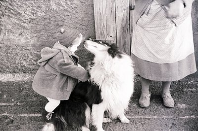 A young child in a hooded coat reaches up to pet a fluffy black and white dog, posed near a weathered wooden door. The woman ...