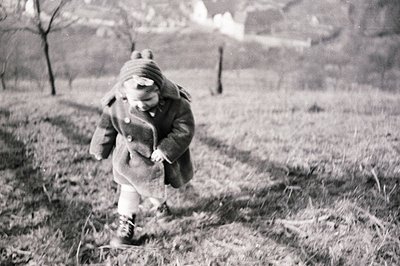 A young child, bundled in a wool coat and hooded hat, runs down a grassy path. The scene appears to be a rural landscape, wit...