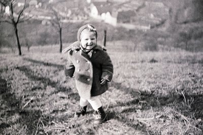 A young child, likely a toddler, runs toward the camera wearing a heavy wool coat, a floral headband, and laced boots. Set ag...