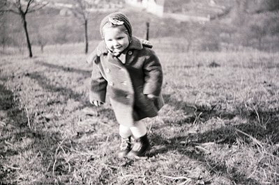 A young child in a wool coat and bonnet runs across a grassy field, captured in a moment of playful motion. The vintage photo...