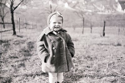 A young child, likely a toddler, stands in a grassy field wearing a heavy wool coat and a decorated hat. The setting appears ...