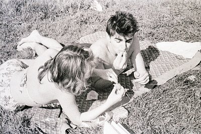 Young couple enjoys a picnic on a grassy lawn. The woman, in a floral print top, offers food to the shirtless man. A patterne...