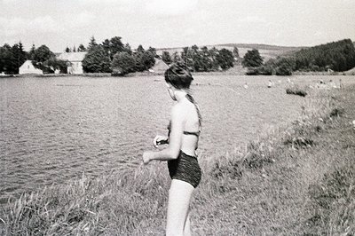 A young woman, viewed from behind, stands near a body of water, likely a lake or reservoir. She wears a textured, dark-colore...