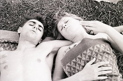 A black and white photo depicts two young people lying closely in grass, seemingly asleep. The male figure is shirtless, and ...
