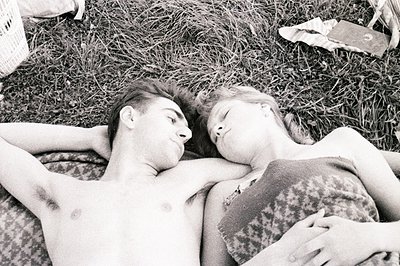 A young couple lies intertwined on a grassy area, heads close together, eyes closed; the man shirtless, the woman on a patter...