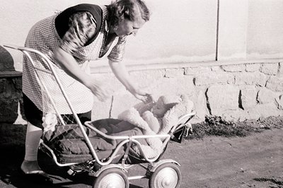 A young woman in a patterned dress and apron tends to a baby in a vintage, wheeled stroller featuring unusual spoke-style whe...