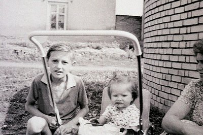 Black and white photo depicts a young boy seated beside a baby in a high-backed stroller. Brick wall and simple architecture ...