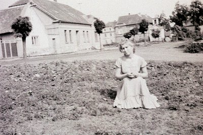 A young woman kneels in a garden, wearing a modest, tiered dress. Background shows residential architecture with multiple hou...