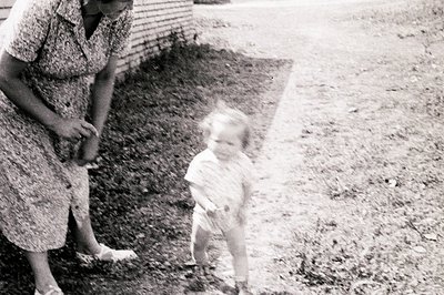 A young child in a diaper and short-sleeved shirt stands on a gravel path, appearing to run towards an adult woman in a patte...