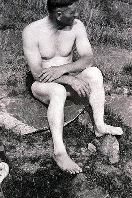 A seated, barefoot man, unclothed from the waist up, rests on a large, textured rock amongst sparse vegetation. The image is ...