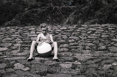 A young girl sits outdoors, cradling a round object (likely a ball) between her legs. She’s barefoot, seated on a grassy area...
