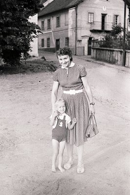 A mother and young child stand on a rural road, framed by a two-story building with a steep, gabled roof. The woman wears a p...