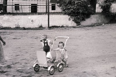 Two young children stand near a vintage metal tricycle; one playfully covers their face. The scene, likely a courtyard or yar...