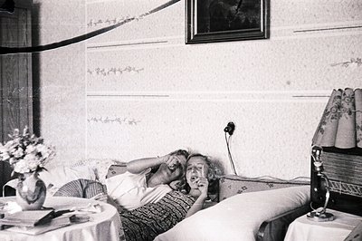 A young woman reclines on a bed, playfully gesturing with one hand near her face. A floral arrangement sits on a nearby table...