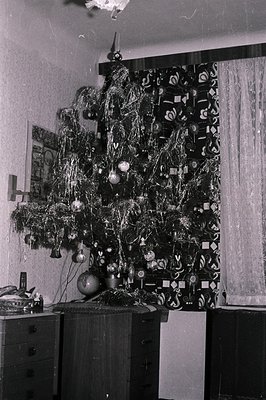 Interior scene showing a tinsel-covered artificial Christmas tree placed in front of a wall with patterned wallpaper and drap...