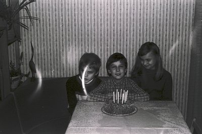 Three children gather around a birthday cake with numerous lit candles on a lace-covered table. Mid-1970s style wallpaper ser...