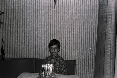 A young boy sits at a table, a birthday cake adorned with numerous lit candles before him. The boy wears a patterned, button-...