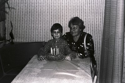 A young boy holds a birthday cake with four candles at a table covered in a lace tablecloth. A smiling elder woman stands bes...