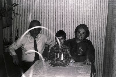 A vintage black and white photo shows a boy celebrating a birthday with his grandparents seated on either side. A cake with l...