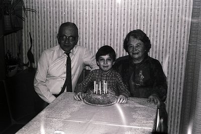 A boy stands proudly with a birthday cake, flanked by an elderly man and woman. The scene evokes a mid-century family celebra...