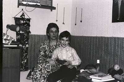 A mother and son pose in a richly decorated room, circa 1970s. The woman wears a patterned dress; the boy, a button-down shir...