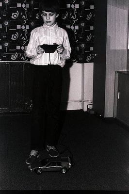 A young boy stands operating a toy radio-controlled car indoors. He wears a button-down shirt and wide-legged trousers, typic...