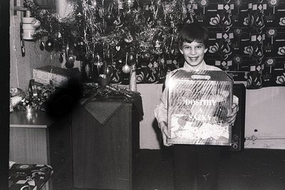 A young boy stands proudly, displaying a "Dostih a Sazky" branded gift box beneath a decorated Christmas tree. Mid-century mo...
