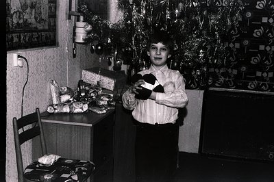 A young boy stands proudly holding a wrapped gift, posed in front of a decorated Christmas tree. Visible are gift packages, a...