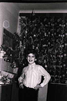 A young boy stands proudly with hands on hips in front of a tinsel-covered Christmas tree. The room’s patterned wallpaper and...