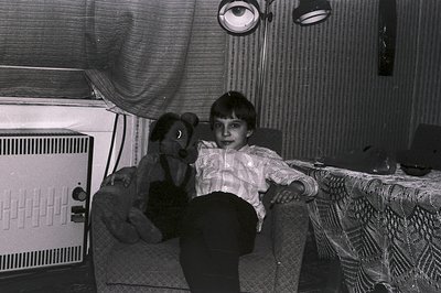 A young boy sits on a patterned armchair, alongside a worn teddy bear. He wears a button-down shirt & dark pants. A textured ...