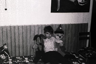 A young boy sits on a patterned sofa, holding a stuffed dog and clown. Visible décor suggests a modest, likely European inter...
