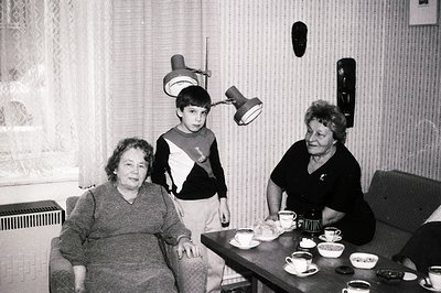 A candid black and white portrait captures a young boy between two women, seemingly at a table set for tea. Architectural det...