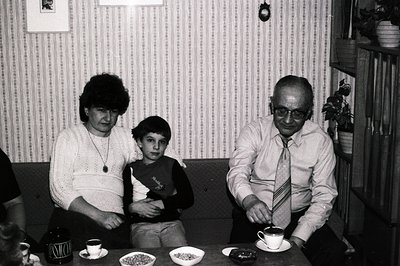 A family portrait, likely 1970s. A grandmother, boy, and grandfather are seated at a table with floral patterned wallpaper in...