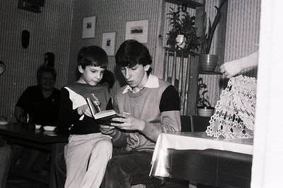 Two boys intently examine a book seated at a table draped with lace. A third person is visible in the background. Likely a do...