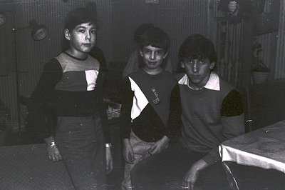 Three young boys in casual, dark-colored sweaters and pants pose in a home interior. A table covered with cloth is visible. L...