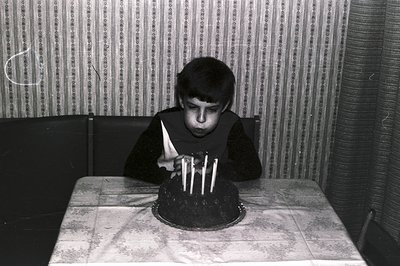 A young boy blows out four candles on a dark chocolate cake covered in white frosting. He holds a folded napkin and is seated...