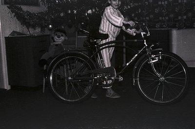 A young boy in striped pajamas stands proudly beside a vintage Schwinn bicycle, likely a gift. A teddy bear sits nearby. Deco...