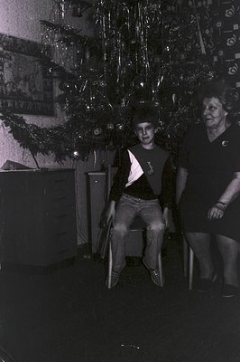 Monochrome portrait of a young man seated under a heavily decorated Christmas tree. An older woman stands to his right, both ...