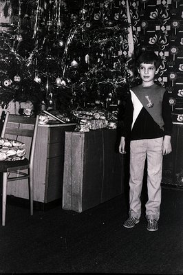 A young boy stands in front of a heavily decorated Christmas tree, surrounded by wrapped gifts. He wears a distinctive color-...