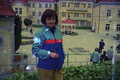A woman in a blue and green tracksuit stands in front of a miniature alpine village, with a fountain in the center and buildi...