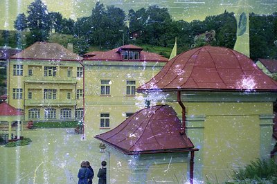 View of a grand, pale yellow building with multiple windows and distinctive red tile rooflines, set within a lush, forested l...