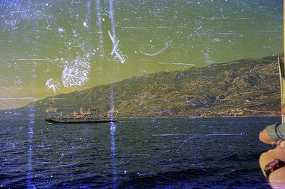 A cargo or ferry ship transits a coastal waterway. Rugged, forested mountains rise in the background with a glimpse of a town...